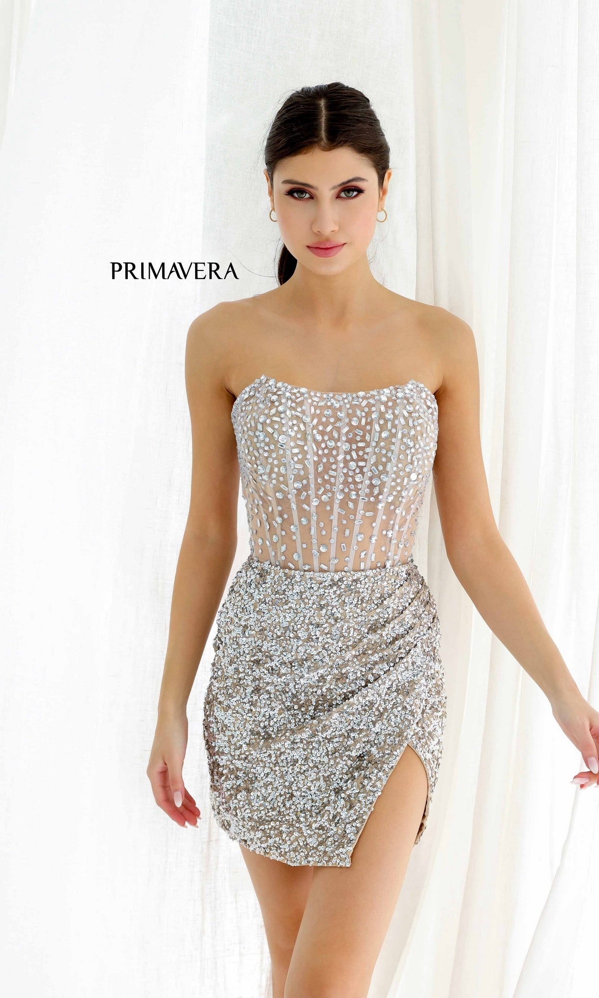 A woman stands in front of white curtains wearing the Short Homecoming Dress 4231 by Primavera, a strapless fitted dress with silver sequins and a side slit. The word